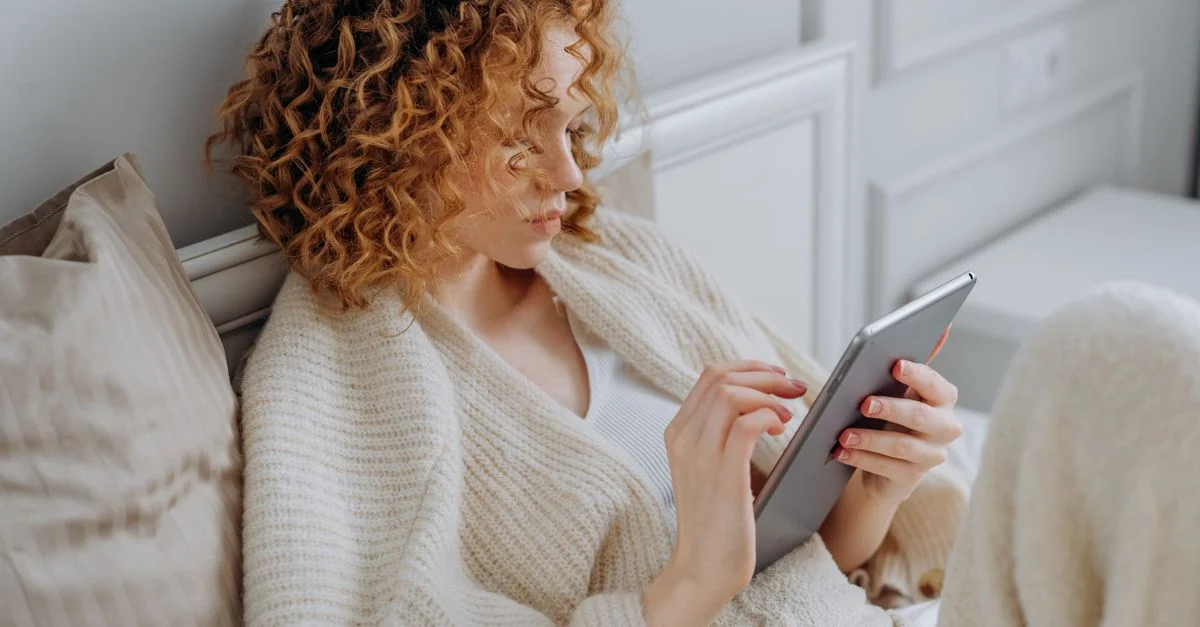 Une femme se détendant sur un lit, utilisant une tablette numérique tout en portant un pull en tricot confortable.